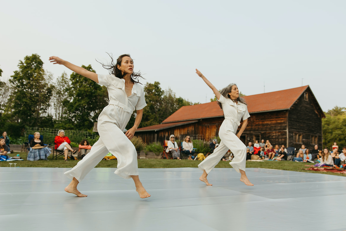 Two dancers in white jumpsuits perform on an outdoor stage with a beautiful, rustic barn behind them.